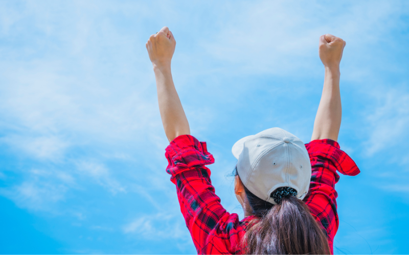 女性が快晴の空に向かって両手を高らかに上げたガッツポーズをしている様子